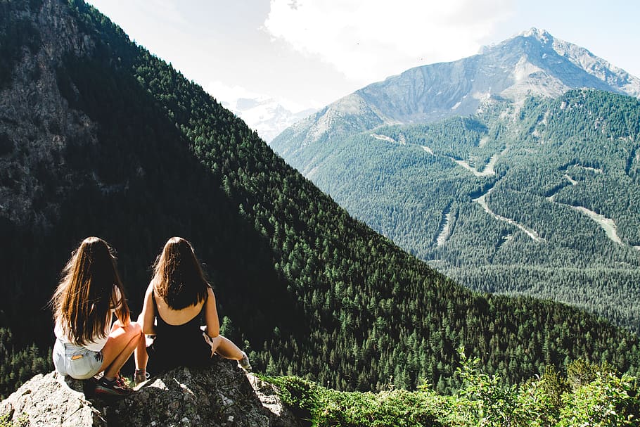 Two Women Sitting on Rock Facing Mountains Under White Cloudy Skies HD wallpaper