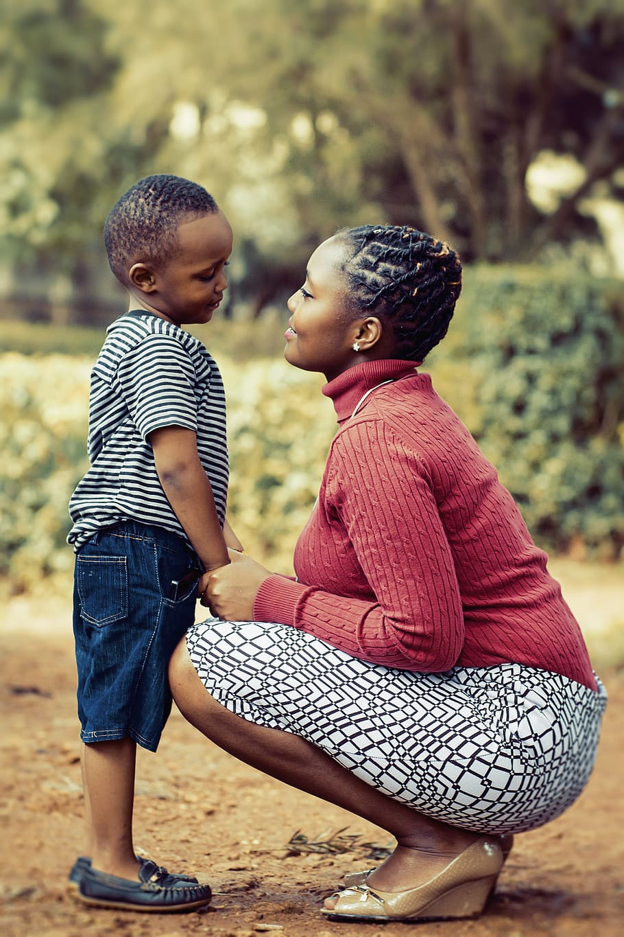 Tilt Shift Lens Photography of Woman Wearing Red Sweater and White Skirt While Holding a Boy Wearing White and Black Crew-neck Shirt and Blue Denim Short