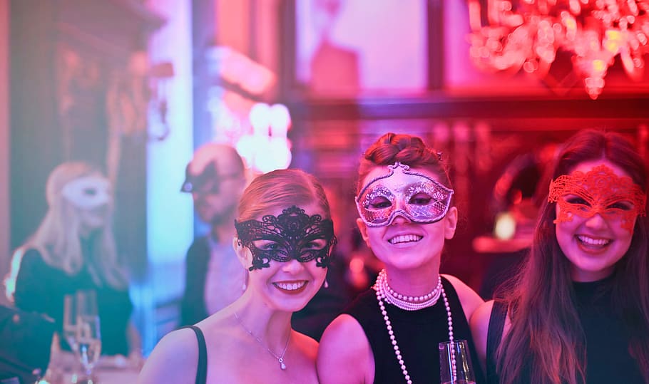 Three Young Women In Masks Posing While Holding Wine Glasses In Nightclub