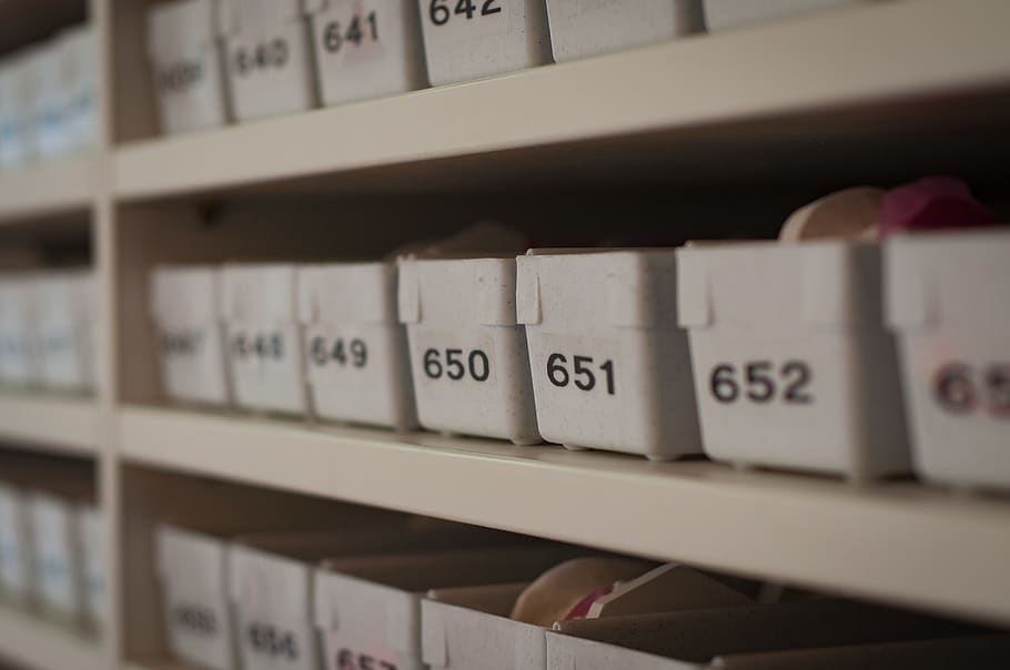  White plastic containers with black labels sit on metal shelves in an organized storeroom.