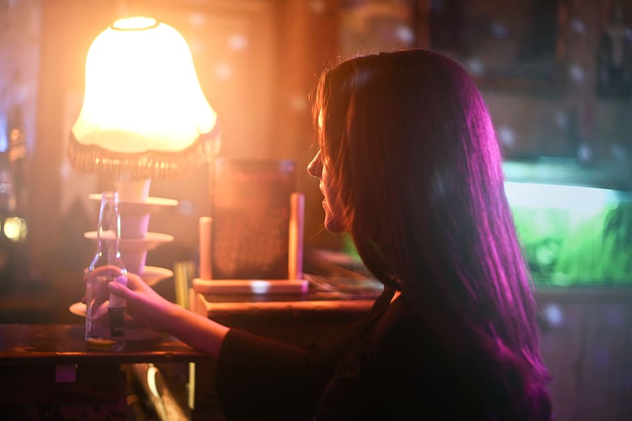 A young brunette woman holding a beer pint bottle in a bar in front of a bright lamp