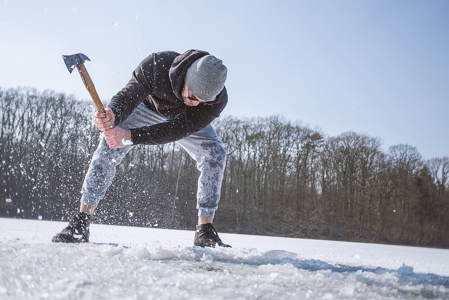 Man Wearing Black Hooded Jacket, Gray Knit Cap, Gray Pants, and Black Shoes Holding Brown Handled Axe While Bending on Snow HD wallpaper