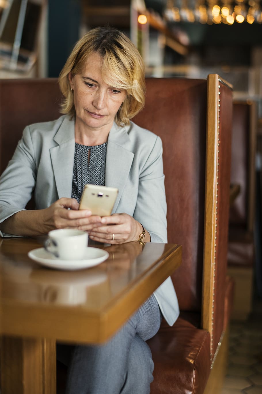 Woman in Grey Notched Lapel Suit Jacket Holding Smartphone Sitting Beside Wooden Table