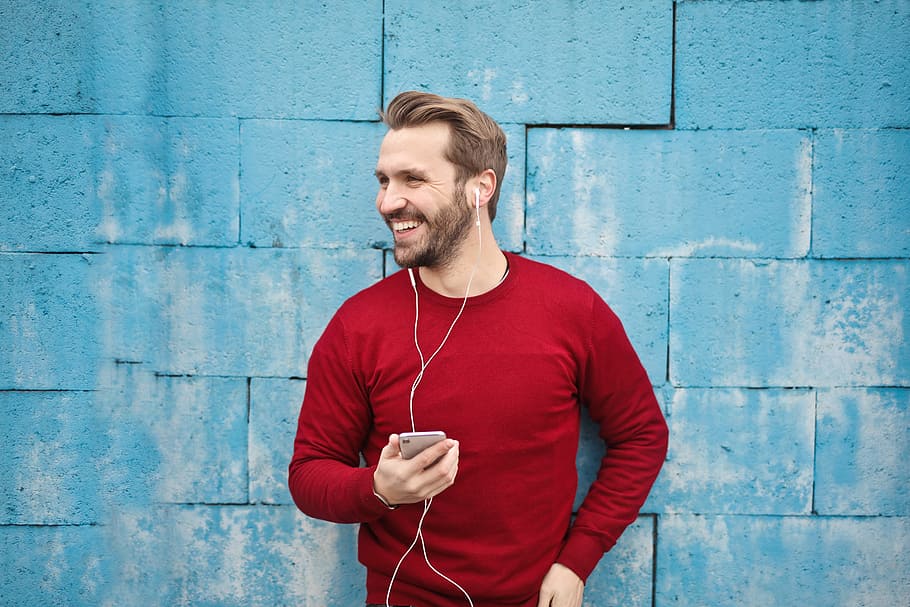 Young Adult Man wearing a red sweater listening to music against turquoise brick wall