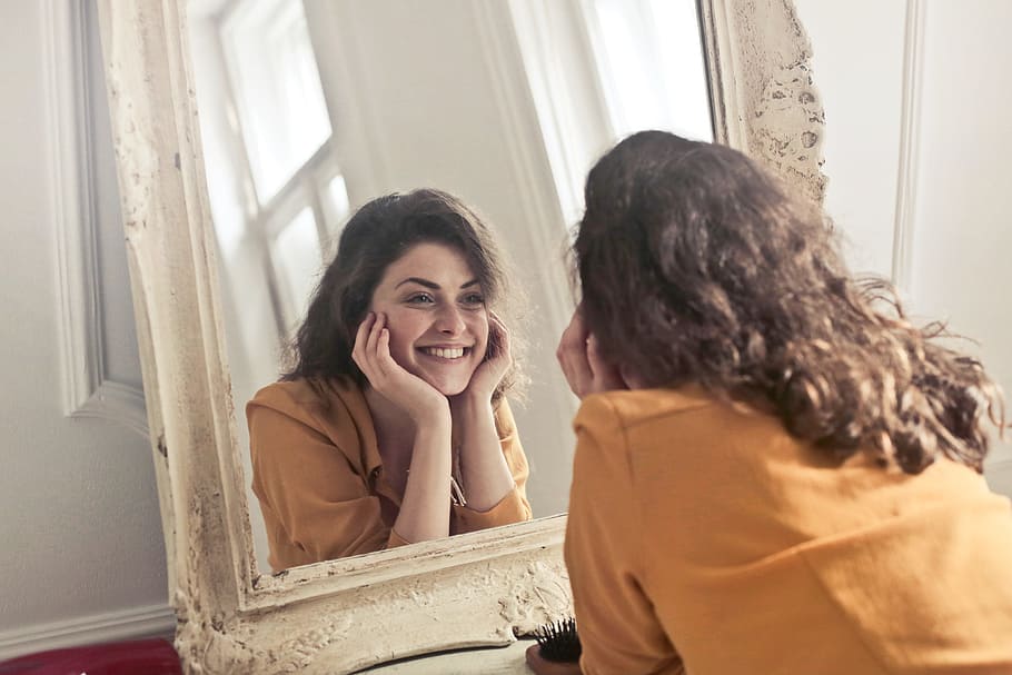 Young woman in Yellow Collared Shirt looking herself reflection in mirror at home