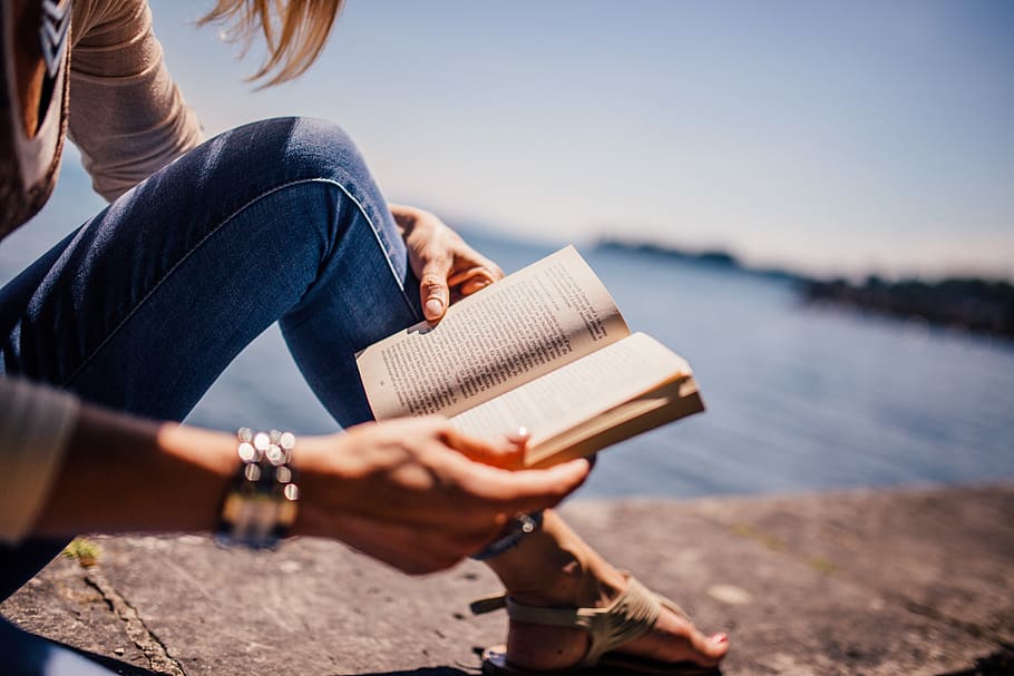 Woman Wearing Blue Denim Jeans Holding Book Sitting on Gray Concrete at Daytime HD wallpaper