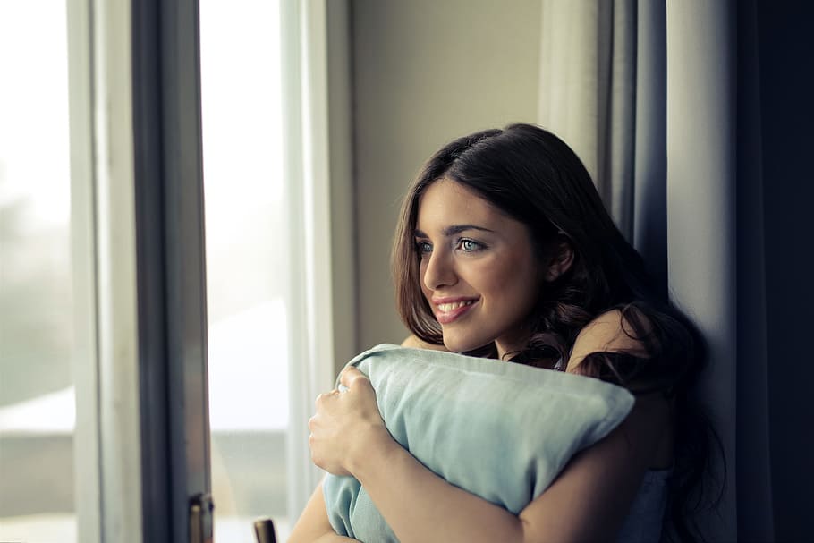 A young brunette woman looks outside the window while emracing a pillow