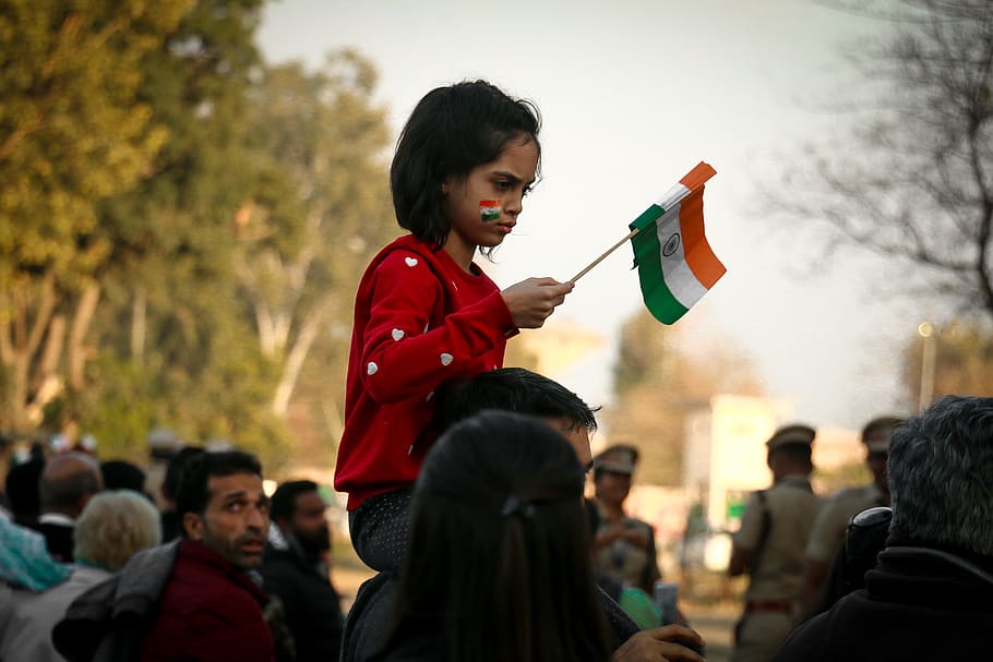 girl sitting on neck of man while holding mini India flag surrounded with people