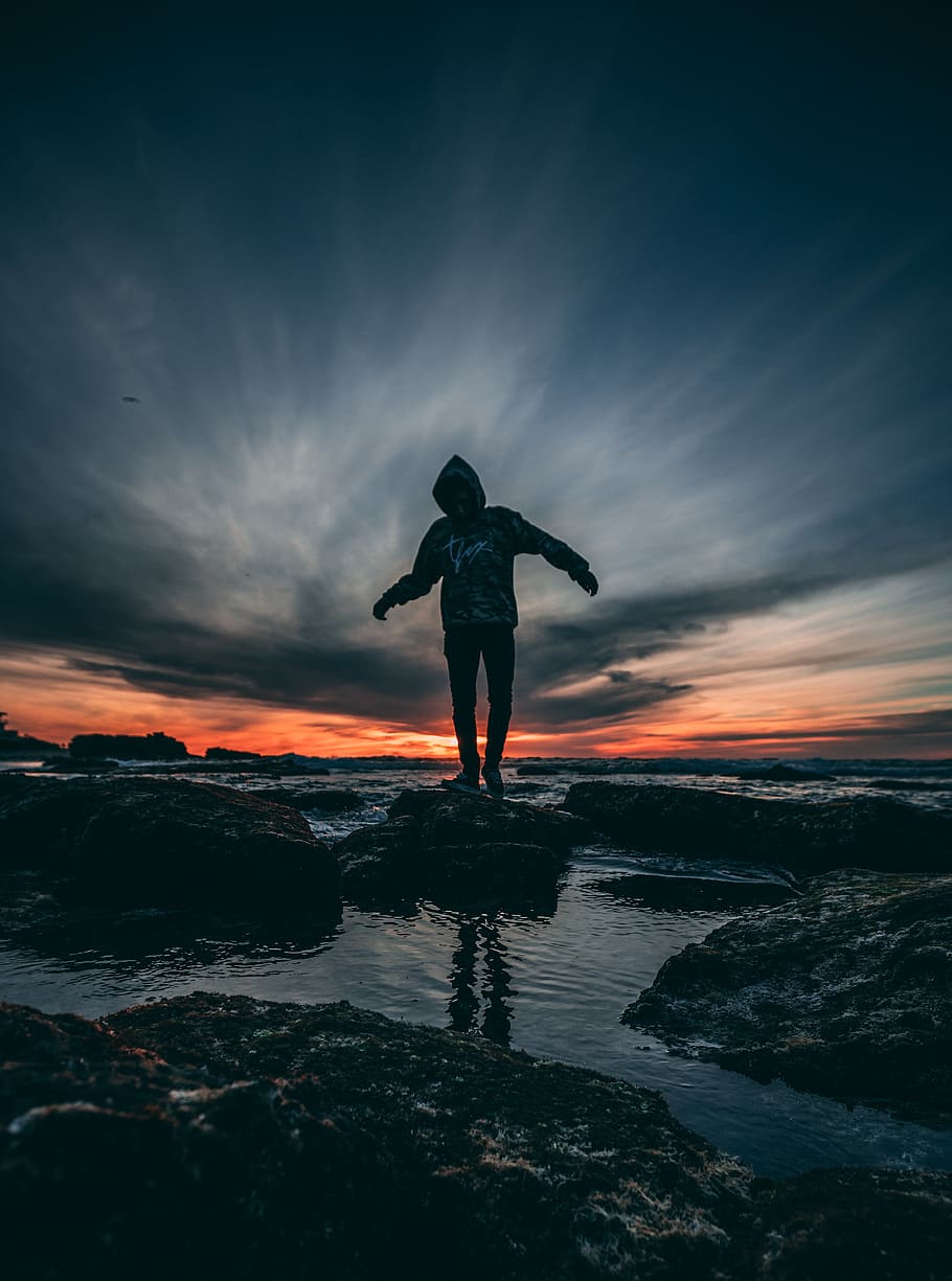 Person Wearing Hoodie Standing on Rock Surrounded by Body of Water HD wallpaper