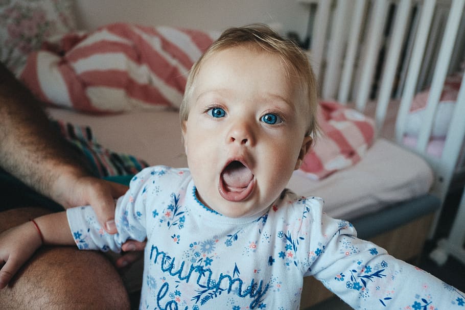 toddler wearing white and blue floral onesie opening mouth wide near white crib