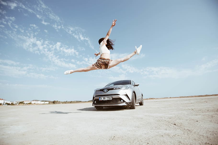 woman jumping in front of parked silver vehicle under clear blue sky during daytime