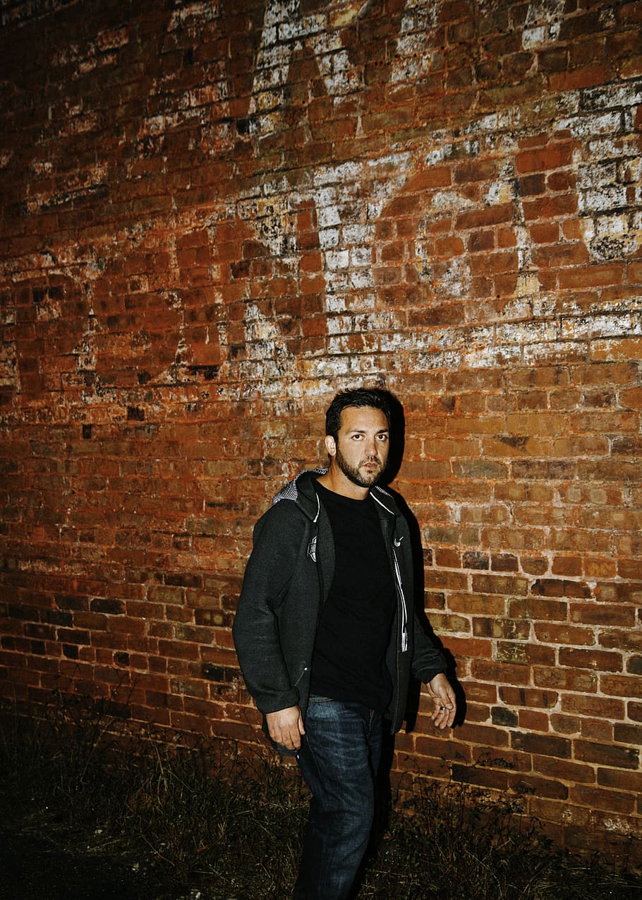 man in black zip-up hoodie walking beside brown bricked wall, HD wallpaper