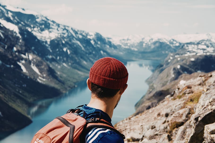 man white red head cap and orange backpack walking along mountain cliff with lake below HD wallpaper