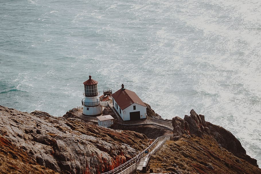 aerial photography of white and brown concrete house beside lighthouse on gray mountain cliff near body of water at daytime HD wallpaper