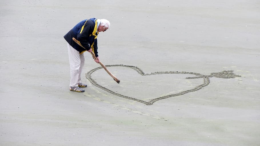 Man Wearing Blue Jacket Holding a Brown Stick Towards the Heart Drawn on Sand HD wallpaper