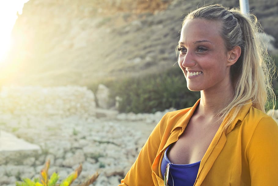 A young blonde woman in yellow button-up shirt and blue tank top posing in the sun with smile