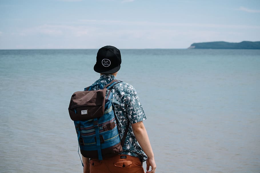 man wearing blue and black backpack standing in front of body of water, HD wallpaper